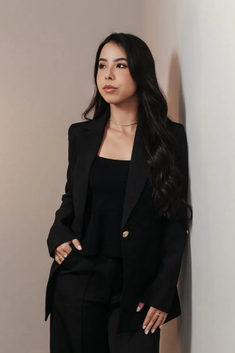 Retrato de Maria Eduarda Coelho, de blazer preto, encostada em uma parede clara
